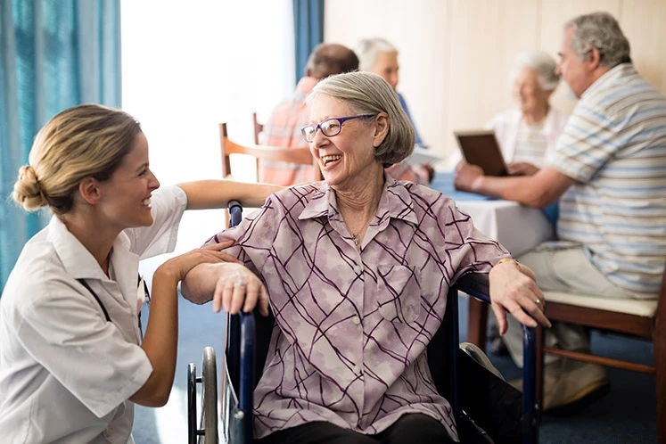 Cheerful disabled senior woman sitting on wheelchair looking at female doctor in retirement home